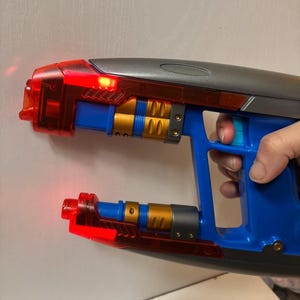 May include: A blue, gray, and gold toy ray gun with red translucent accents. The toy has a trigger and a handle, and the barrel has a red light. The toy is held by a person.