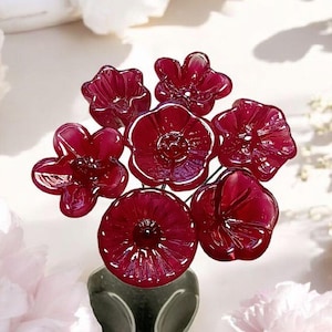 May include: A bouquet of seven burgundy glass flowers in a black vase. The flowers are arranged in a cluster and have a delicate, intricate design.