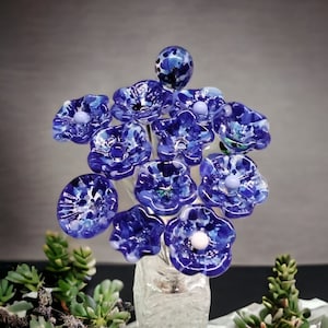 May include: A bouquet of blue glass flowers with white speckles, arranged in a clear glass vase. The flowers are made of glass and have a delicate, whimsical appearance.