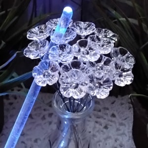 May include: A clear glass vase filled with a bouquet of clear glass flowers. The flowers are lit from within by a blue light source, creating a glowing effect.
