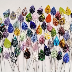 May include: A collection of colorful glass leaf-shaped ornaments on wire stems. The leaves are in various colors, including blue, green, yellow, and purple, with intricate vein details. These decorative pieces are likely for floral arrangements or crafts.