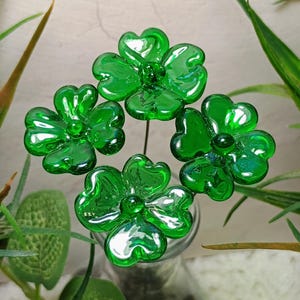 May include: Four green glass shamrock flowers with a shiny finish are arranged in a vase. The flowers are made of glass and have a realistic appearance.