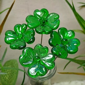 May include: Four green glass flowers with four-leaf clover designs are arranged in a vase. The flowers are made of clear glass with a green tint.
