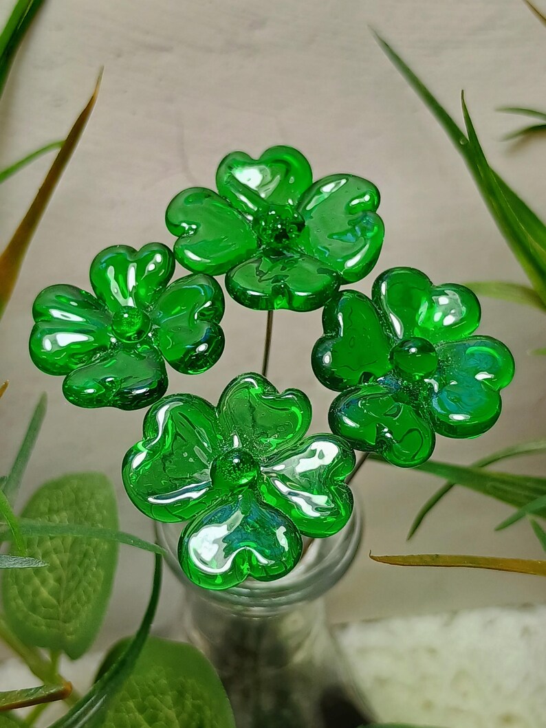 May include: Four green glass flowers with four-leaf clover designs are arranged in a vase. The flowers are made of clear glass with a green tint.