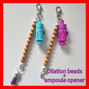 May include: Two keychains with syringe charms in blue and purple, each with a smiling face. They hang from wooden bead strands and silver-tone hardware. The text "Dilation beads + ampoule opener" is visible.