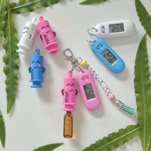 May include: A collection of syringe-shaped charms in white, pink, and blue, alongside digital watch fobs in white and blue. One pink syringe charm is attached to a pink digital watch fob and a beaded keychain spelling the name CHARLOTTE.