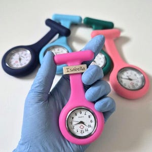 May include: A selection of colourful nurse fob watches in pink, blue, green, and navy. The pink watch, held by a gloved hand, has the name "Isabella" on it. Each watch features a white clock face with black numerals and hands.