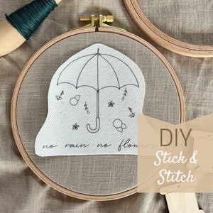 Könnte beinhalten: Ein DIY-Stickset mit einem weißen Stoffring, auf dem eine schwarze Linienzeichnung eines Regenschirms und der Text "no rain no flowers" zu sehen ist. Das Set enthält einen Stickrahmen aus Holz, eine Spule mit grünem Stickgarn und eine Holzspule.