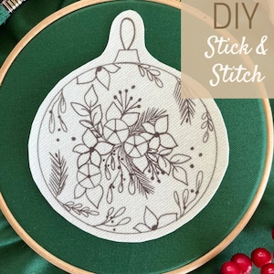 Può includere: Un ornamento in tessuto bianco con un motivo floreale nero in un telaio da ricamo in legno. Il telaio è su una superficie di tessuto verde. Il testo "DIY Stick & Stitch" è nell'angolo in alto a destra dell'immagine.