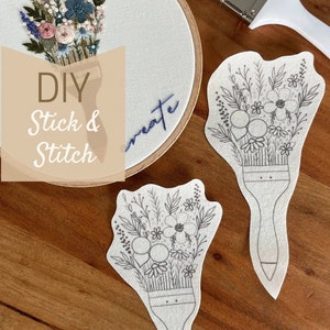 Könnte beinhalten: DIY Stick & Stitch Stickerei-Projekt mit einem floralen Design auf weißem Stoff. Das Projekt beinhaltet ein vorgefertigtes Design auf weißem Stoff, einen hölzernen Stickrahmen und einen Pinsel.