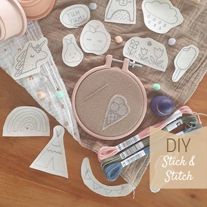 Könnte beinhalten: Ein DIY-Stickset mit einer Vielzahl süßer Designs, darunter ein Einhorn, ein Bär, ein Regenbogen, ein Tipi, eine Wolke, eine Birne, ein Apfel, eine Blume, ein Eiswaffel und ein Eis am Stiel. Das Set enthält Stickgarn, eine Nadel und einen Stickrahmen. Der Text "DIY Stick & Stitch" ist auf dem Bild gedruckt.