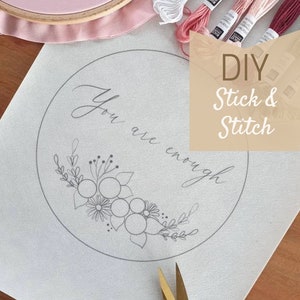 Könnte beinhalten: Ein DIY-Stickset mit einer weißen Leinwand, einem Stickrahmen und Stickgarn. Die Leinwand hat ein Design mit dem Text "You are enough" und einem floralen Muster. Das Set enthält außerdem eine goldene und schwarze Stickerschere.