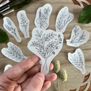 May include: A set of nine white fabric patches with black line drawings of various floral designs. The patches are on a wooden surface with green embroidery floss.