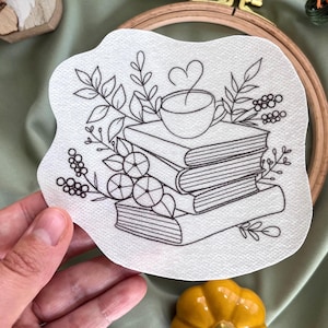 Könnte beinhalten: Eine Schwarzweiß-Zeichnung eines Stapels Bücher mit einer Tasse Kaffee darauf. Die Bücher sind von Blättern und Blumen umgeben.