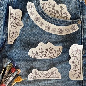 Könnte beinhalten: Nahaufnahme von Jeansshorts mit floralen Stickereien. Die Shorts haben eine klassische blaue Waschung und verfügen über mehrere gestickte Aufnäher mit zarten Blumen- und Blättermustern. Stickgarn in verschiedenen Farben ist sichtbar.