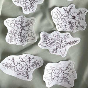 Peut inclure: Six motifs de broderie florale noirs et blancs sur tissu blanc. Les motifs sont disposés en demi-cercle sur un fond de tissu vert. Un cerceau de broderie en bois se trouve dans le coin inférieur gauche de l'image.