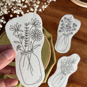 Könnte beinhalten: Drei weiße Stoff-Stickmuster von Blumen in einem Einmachglas. Die Muster sind in Form des Glases und der Blumen ausgeschnitten.