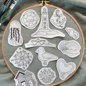 Könnte beinhalten: Ein Set mit 12 schwarzen und weißen Stickmustern auf weißem Papier mit nautischen Motiven wie einem Leuchtturm, Anker, Kompass, Muscheln und einem Segelboot. Die Muster sind in einem Kreis auf einem hölzernen Stickrahmen angeordnet.