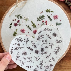 Könnte beinhalten: Ein weißes herzförmiges Stück Stoff mit einem braunen Blumenmuster wird vor einem weißen Stickrahmen mit einem braunen Blumendesign gehalten. Der Rahmen ist mit einer braunen Schnur gebunden.