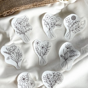 Könnte beinhalten: Sieben schwarz-weiße Stickmuster mit verschiedenen Blumensträußen auf weißem Stoff.