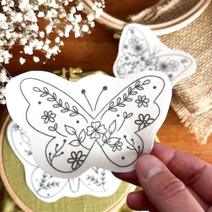 Könnte beinhalten: Ein weißes Stoff-Schmetterlings-Stickmuster mit floralem Design. Der Schmetterling ist mit schwarzem Garn umrandet und hat ein zartes Blumenmuster.