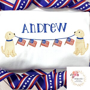 Op de afbeelding: Witte geborduurde T-shirt met de naam "Andrew" en twee gele honden die een string Amerikaanse vlaggen vasthouden.