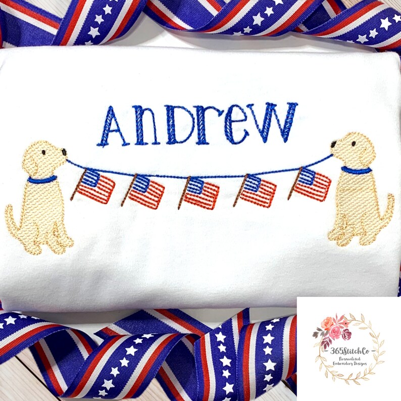 Op de afbeelding: Witte geborduurde T-shirt met de naam "Andrew" en twee gele honden die een string Amerikaanse vlaggen vasthouden.