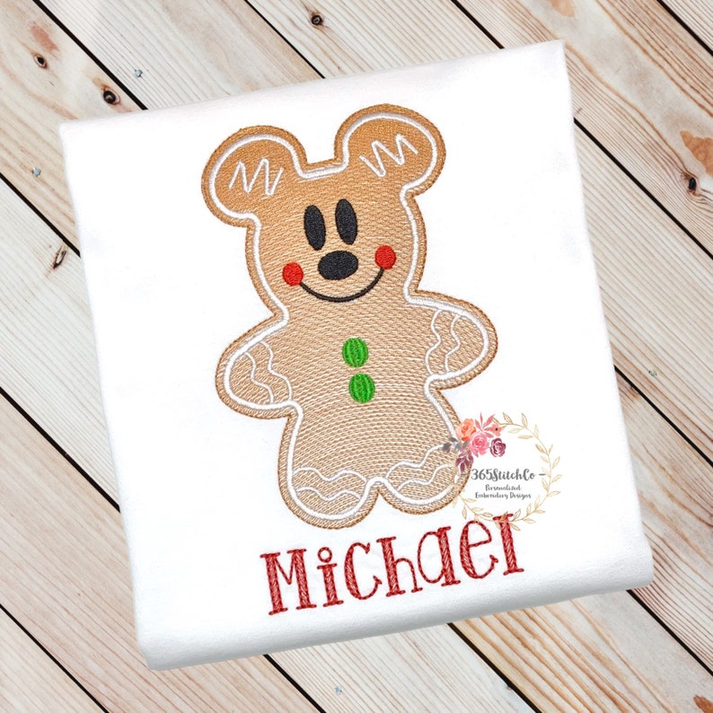 May include: A gingerbread man with Mickey Mouse ears and a red smile, embroidered on a white shirt with the name "Michael" in red lettering.