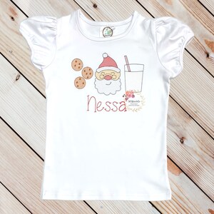 May include: White short-sleeved t-shirt with a red and white Santa Claus face, three brown cookies, and a glass of milk with a red straw. The name "Nessa" is embroidered in red below the image.