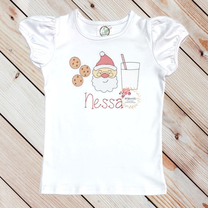 May include: White short-sleeved t-shirt with a red and white Santa Claus face, three brown cookies, and a glass of milk with a red straw. The name "Nessa" is embroidered in red below the image.