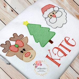 Puede incluir: Un pelele blanco con un reno bordado en rojo, blanco y marr&oacute;n, un &aacute;rbol de Navidad verde y un Pap&aacute; Noel rojo y blanco. El pelele tambi&eacute;n tiene el nombre "Kate" bordado en rojo.