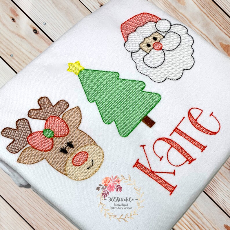 Puede incluir: Un pelele blanco con un reno bordado en rojo, blanco y marr&oacute;n, un &aacute;rbol de Navidad verde y un Pap&aacute; Noel rojo y blanco. El pelele tambi&eacute;n tiene el nombre "Kate" bordado en rojo.