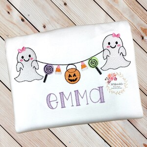 Puede incluir: Camiseta blanca con un nombre bordado en morado "EMMA" y un dise&ntilde;o de tem&aacute;tica de Halloween con dos fantasmas con lazos rosas, ma&iacute;z dulce, piruletas y una calabaza de Halloween.