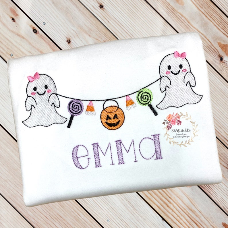 Puede incluir: Camiseta blanca con un nombre bordado en morado "EMMA" y un dise&ntilde;o de tem&aacute;tica de Halloween con dos fantasmas con lazos rosas, ma&iacute;z dulce, piruletas y una calabaza de Halloween.