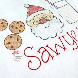 May include: Embroidered design featuring a cartoon Santa Claus with a glass of milk and cookies. The text "Sawyer" is embroidered in red below the design.