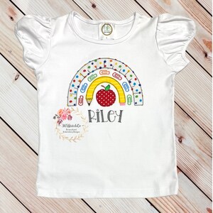 May include: White short-sleeved shirt with a rainbow design made of school supplies, including pencils, paper clips, and a red apple. The name "Riley" is embroidered below the rainbow.