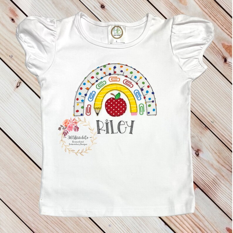 May include: White short-sleeved shirt with a rainbow design made of school supplies, including pencils, paper clips, and a red apple. The name "Riley" is embroidered below the rainbow.