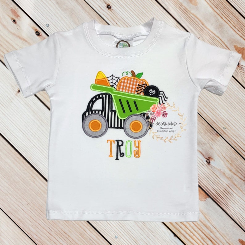 Puede incluir: Camiseta blanca de manga corta con un cami&oacute;n verde que lleva una calabaza, una ara&ntilde;a y ma&iacute;z dulce. El nombre "TROY" est&aacute; bordado en verde debajo del cami&oacute;n.