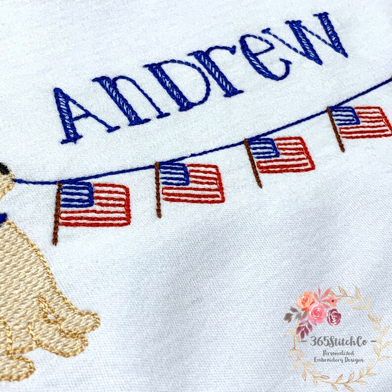 Op de afbeelding: Een geborduurd ontwerp met de naam "Andrew" in blauwe draad, met daaronder een rij Amerikaanse vlaggen in rood, wit en blauw. Links van het ontwerp is een kleine geborduurde hond te zien.