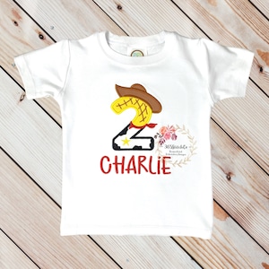 K&ouml;nnte beinhalten: Wei&szlig;es T-Shirt mit einer roten, gelben und braunen gestickten Zahl Zwei mit einem Cowboyhut. Die Zahl Zwei hat einen schwarz-wei&szlig;en Kuhmuster-Hintergrund. Der Name "CHARLIE" ist in Rot unter der Zahl gestickt.
