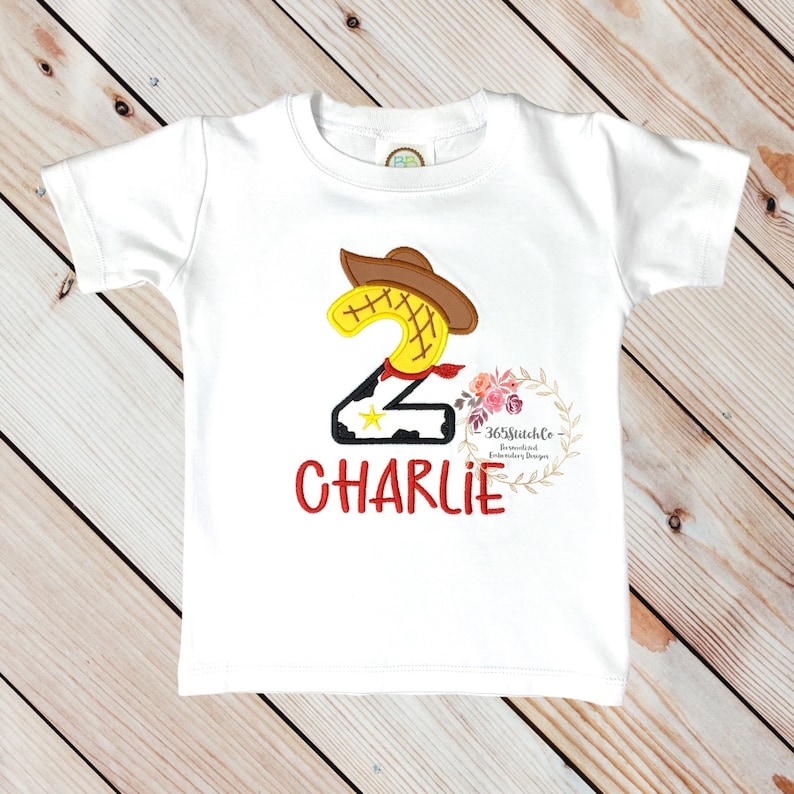 K&ouml;nnte beinhalten: Wei&szlig;es T-Shirt mit einer roten, gelben und braunen gestickten Zahl Zwei mit einem Cowboyhut. Die Zahl Zwei hat einen schwarz-wei&szlig;en Kuhmuster-Hintergrund. Der Name "CHARLIE" ist in Rot unter der Zahl gestickt.