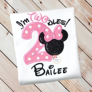 Puede incluir: Una camiseta blanca con bordados en rosa y negro. El n&uacute;mero "2" es rosa con lunares blancos y tiene un lazo rosa en la parte superior. El texto "I'm Two-dles!" est&aacute; en cursiva negra. El nombre "Bailee" est&aacute; en cursiva negra debajo del n&uacute;mero.