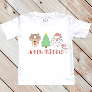May include: White t-shirt with an embroidered design of a reindeer, Christmas tree, and Santa Claus in red, green, and brown. The text "Jorge Alberto" is embroidered in red below the design.