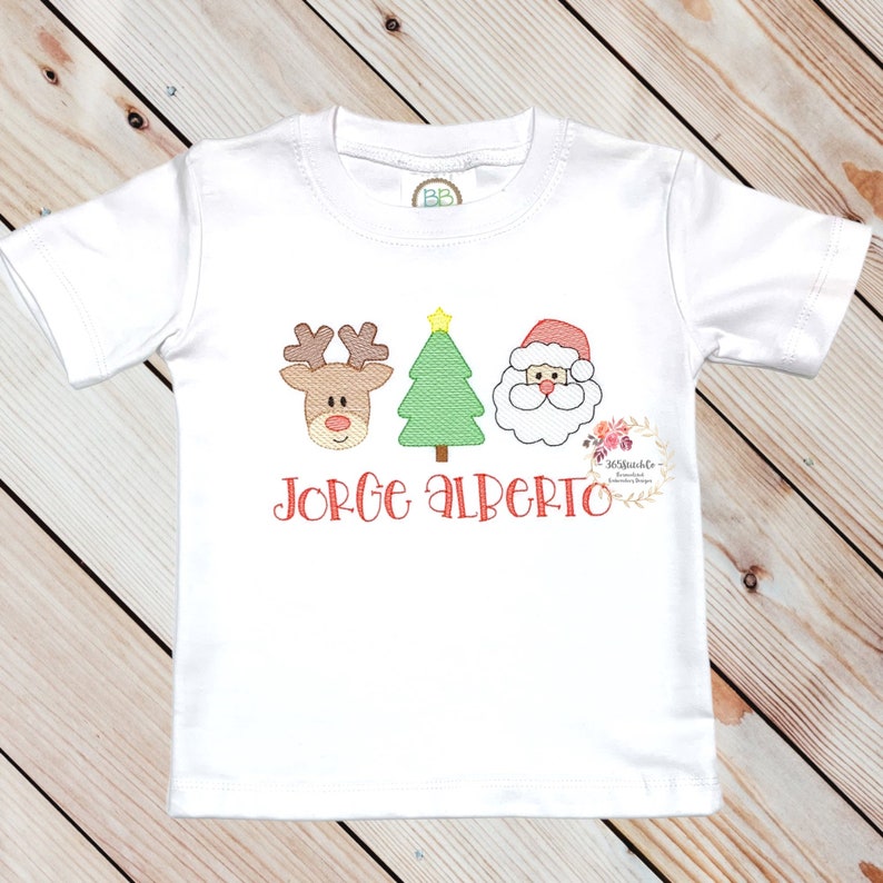 May include: White t-shirt with an embroidered design of a reindeer, Christmas tree, and Santa Claus in red, green, and brown. The text "Jorge Alberto" is embroidered in red below the design.
