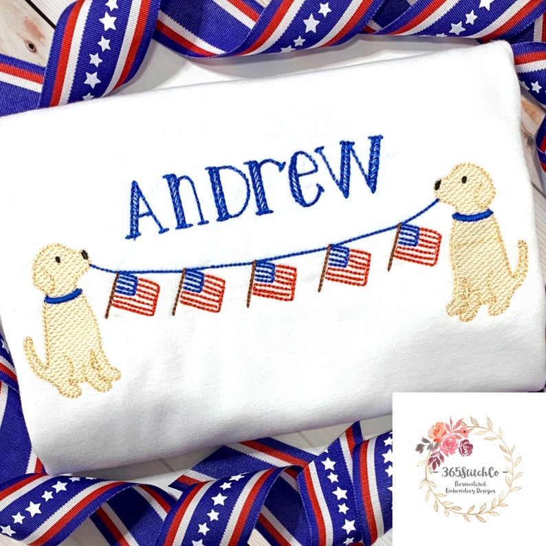 Op de afbeelding: Witte geborduurde t-shirt met de naam "Andrew" en twee gele honden die een touwtje met Amerikaanse vlaggen vasthouden.