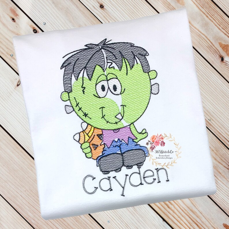 May include: A white t-shirt with an embroidered design of a green Frankenstein monster holding a candy corn. The monster has black hair, a purple shirt, blue jeans, and black shoes. The name "Cayden" is embroidered below the monster.
