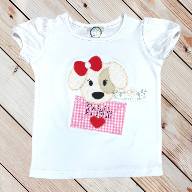 May include: White t-shirt with a red and white gingham envelope with a heart and the name "Brielle" embroidered on it. A brown and white dog with a red bow is peeking out of the envelope.