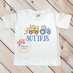 May include: White short-sleeved t-shirt with an embroidered design of a blue tractor pulling a wagon full of carrots. A bunny is driving the tractor. The name "SOTIRIS" is embroidered below the tractor. The text "365StitchCo Personalized Embroidery Designs" is embroidered in a wreath at the bottom of the shirt.