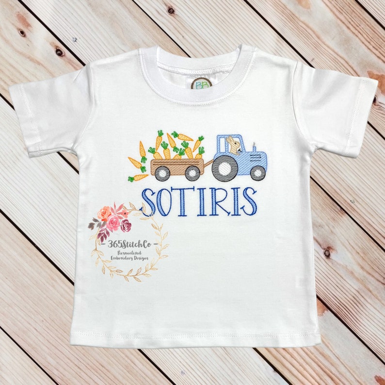 May include: White short-sleeved t-shirt with an embroidered design of a blue tractor pulling a wagon full of carrots. A bunny is driving the tractor. The name "SOTIRIS" is embroidered below the tractor. The text "365StitchCo Personalized Embroidery Designs" is embroidered in a wreath at the bottom of the shirt.