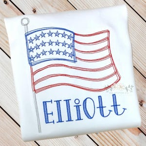 K&ouml;nnte beinhalten: Wei&szlig;es T-Shirt mit einer gestickten amerikanischen Flagge und dem Namen "Elliott" in blauer Farbe.